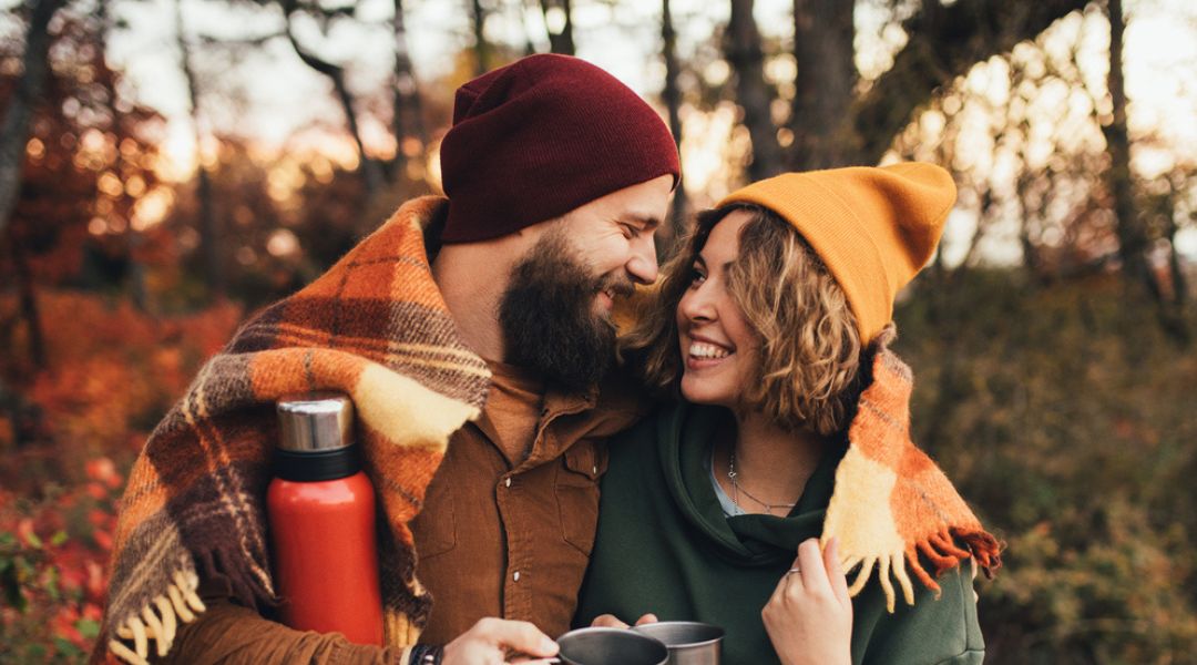 Smiling couple holding camping mugs in the woods, autumn.